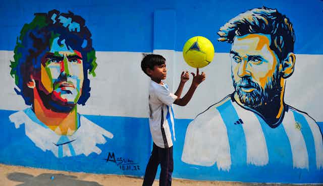 A boy balances a yellow soccer ball on his finger in front of a mural showing Diego Maradona and Lionel Messi.