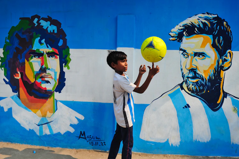 A boy balances a yellow soccer ball on his finger in front of a mural showing Diego Maradona and Lionel Messi.