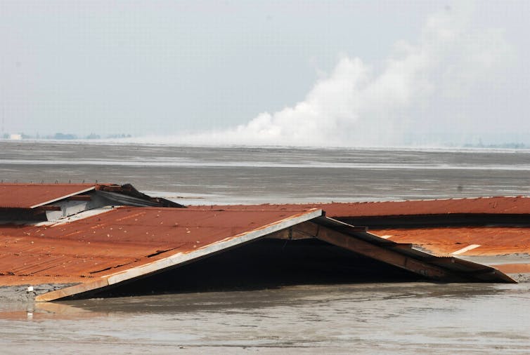 Houses are submerged in mud, while gas billows from mud volcano in background.