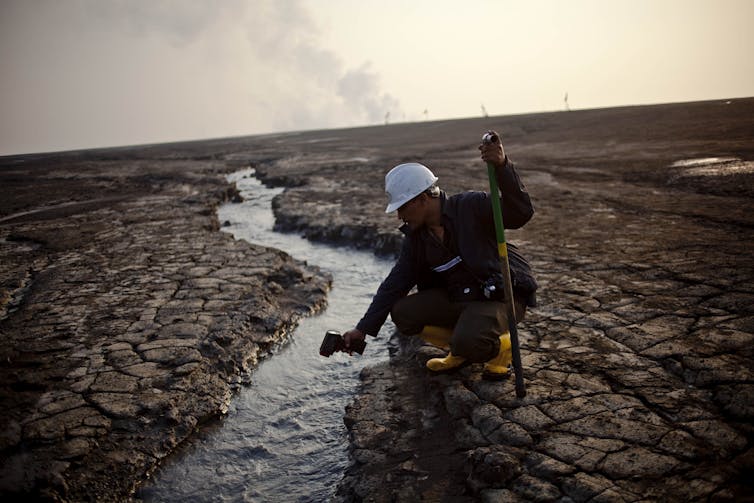 Man kneels on cracking dry mud points thermometer at a flowing stream