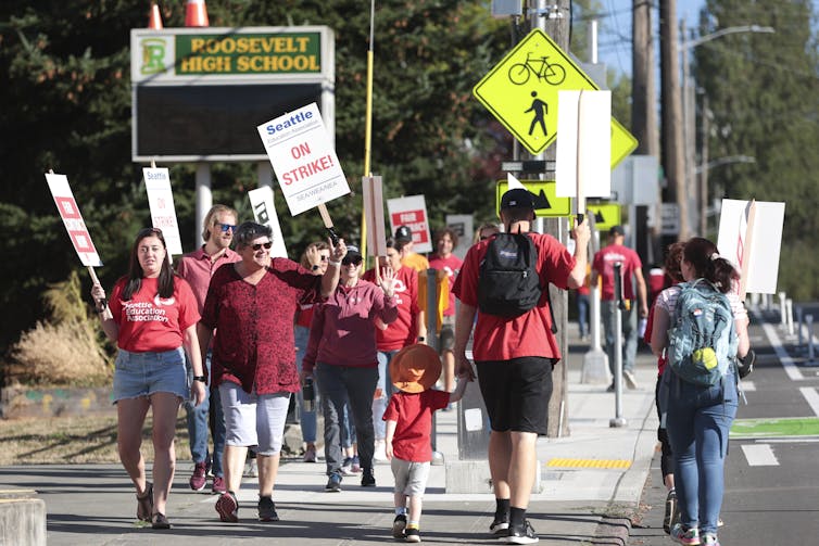 Striking education workers seen with picket signs walking on a sidewalk.