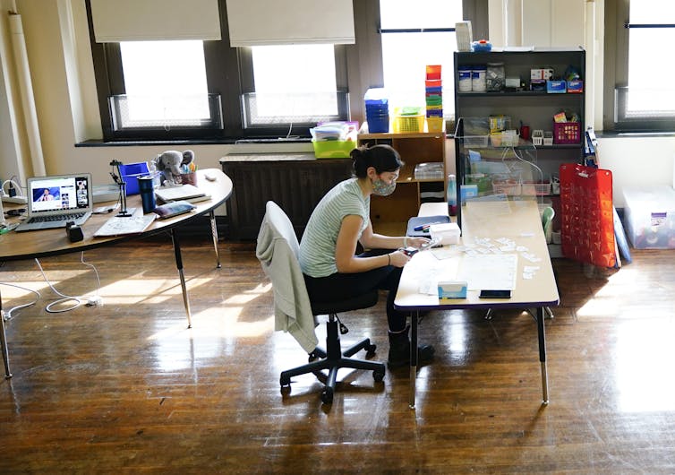 A teacher seen sitting at a desk cutting things with a laptop behind her on a desk.