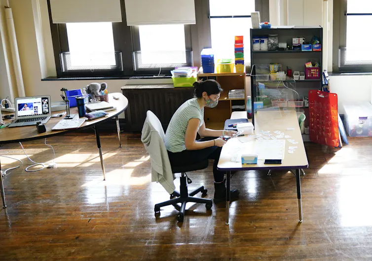 A teacher seen sitting at a desk cutting things with a laptop behind her on a desk.