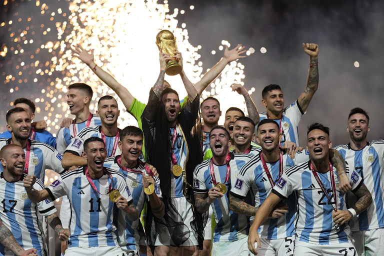 Football players raise their arms in celebration. One of them is holding a golden trophy.