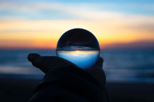 A photo of a hand holding up a crystal ball in front of a beach sunrise.