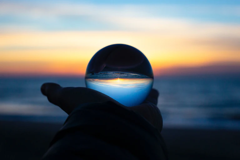 A photo of a hand holding up a crystal ball in front of a beach sunrise.