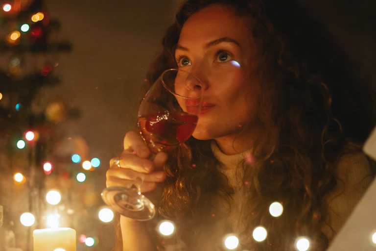 A curly-haired woman drinking alone near a Christmas tree, candles and lights