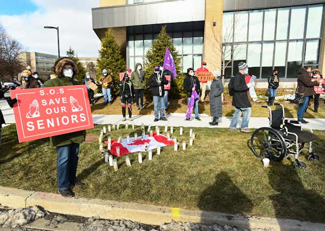 A group of protesters in the background; in the foreground a person holds a sign reading 'S.O.S. save our seniors'