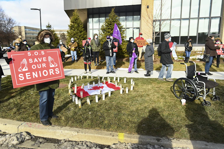 A group of protesters in the background; in the foreground a person holds a sign reading 'S.O.S. save our seniors'