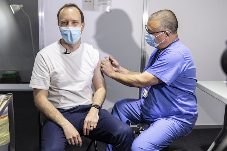 Hancock smiles wearing a mask and a white t-shirt as he receives his COVID vaccination.