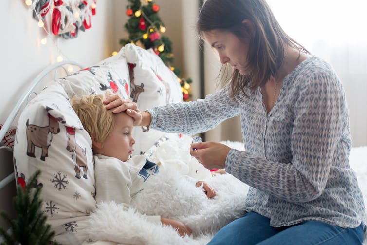 A mother feels her child's forehead. The child is in bed and the bedroom is decorated for Christmas.