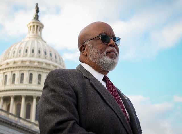 A gray-bearded black man dressed in a dark business suit stands in front of a building that has a white dome.