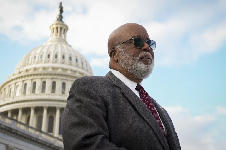 A gray-bearded black man dressed in a dark business suit stands in front of a building that has a white dome.