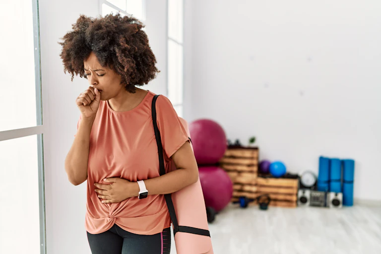 A woman with a yoga mat on her shoulder coughs into her fist.