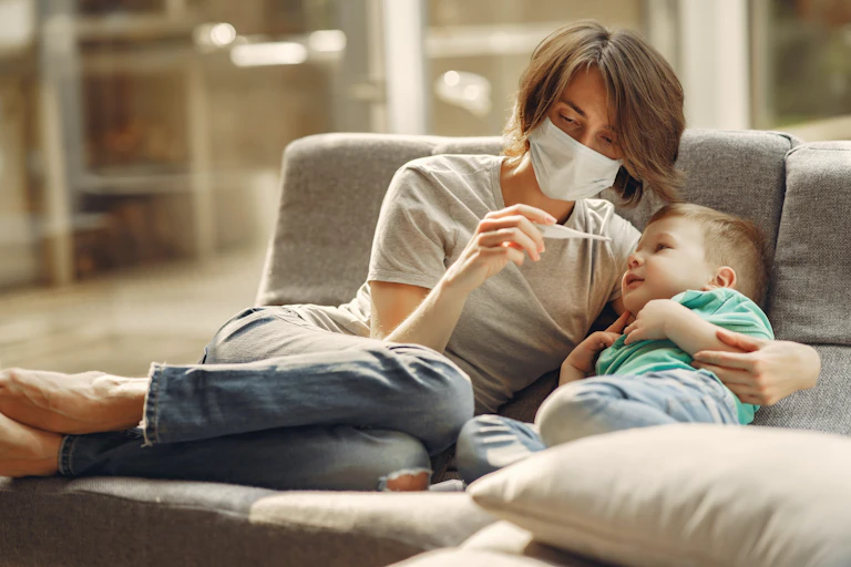 A woman in a face mask holding a child on a sofa, looking at a thermometer