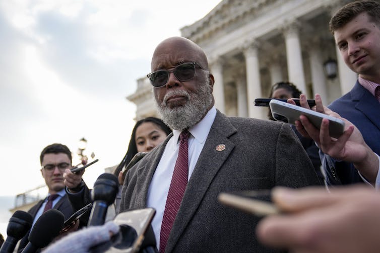 A middle aged Black man with a white beard wears sunglasses and stands in front of the U.S. Capitol building, as he is surrounded by people holding up voice recorders and phones.