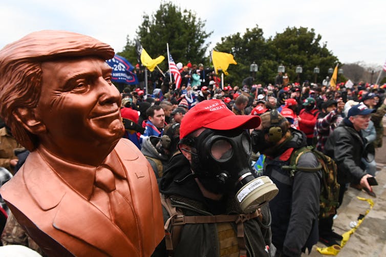 A man with a gas mask stands in a crowd of people with Trump and American flags and holds up a bronze bust of a man wearing a suit