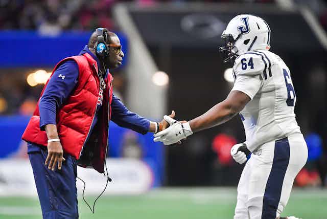 Coach wearing headphones congratulates football player in uniform.