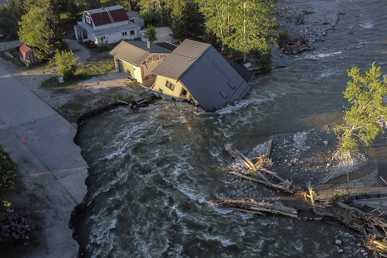 A house is tipped into the river after water cut a new channel beneath it. It also tore out a road and bridge.