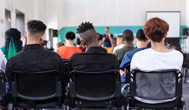 Three people are photographed from behind in a university classroom, one with short dreadlocks.
