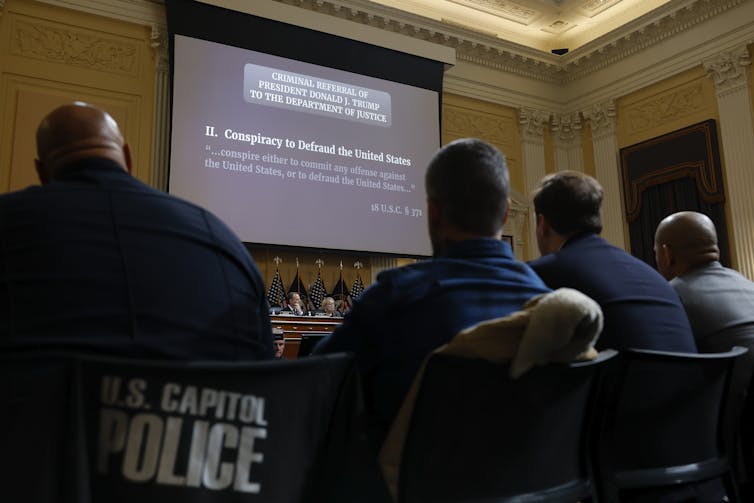 The backs of four people are seen looking at a projector screen that says 'Two. Conspiracy to defraud the United States,' with the words 'criminal referral of President Donald J. Trump to the Department of Justice.'