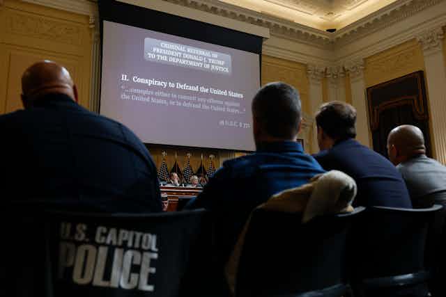 The backs of four people are seen looking at a projector screen that says "Two. Conspiracy to defraud the United States," with the words "criminal referral of President Donald J. Trump to the Department of Justice."