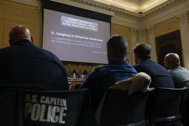 The backs of four people are seen looking at a projector screen that says 'Two. Conspiracy to defraud the United States,' with the words 'criminal referral of President Donald J. Trump to the Department of Justice.'
