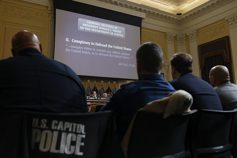 The backs of four people are seen looking at a projector screen that says "Two. Conspiracy to defraud the United States," with the words "criminal referral of President Donald J. Trump to the Department of Justice."