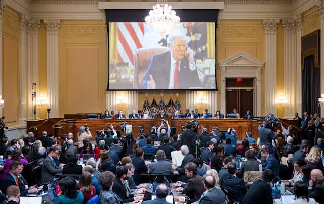 A big screen shows a man in a blue suit and red tie gesticulating while on the phone. Below him sit a panel of people and a bank of photographers.