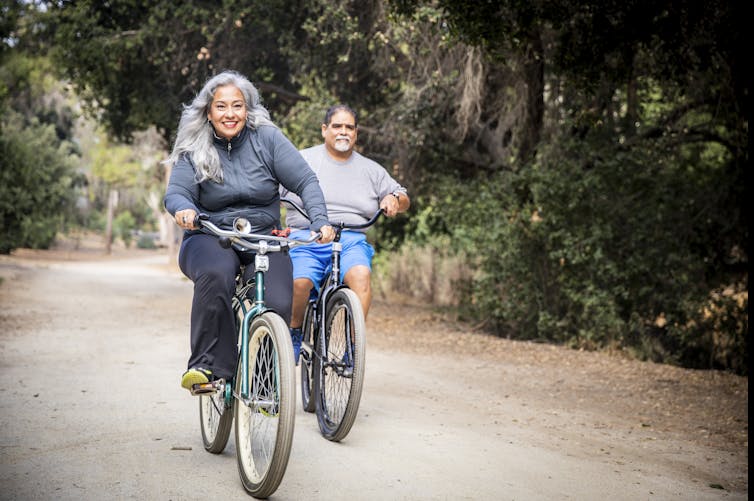 Middle-aged woman with long gray hair riding a bicycle in foreground and middle-aged man riding a bike in background.
