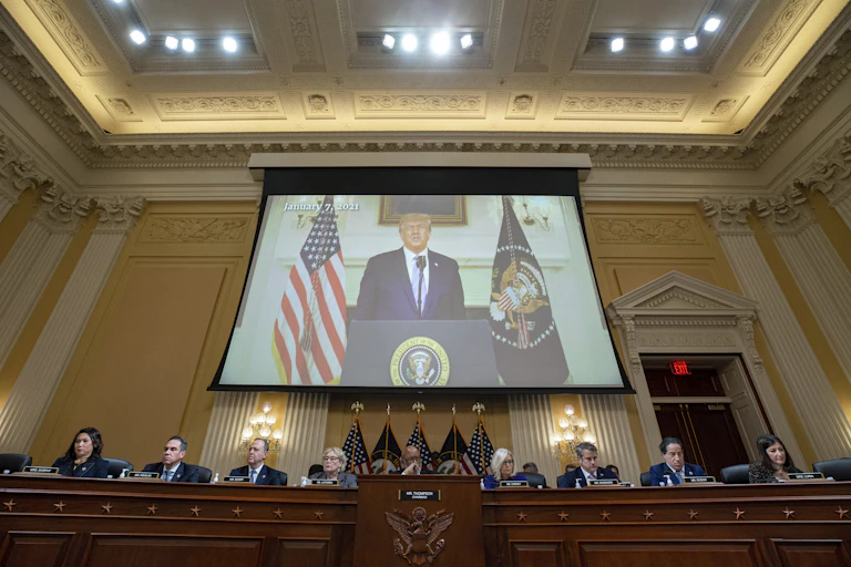 A screen shows a man in a suit between a U.S. flag and a flag bearing the seal of the U.S. president, high above a row of people seated at a wooden desk.