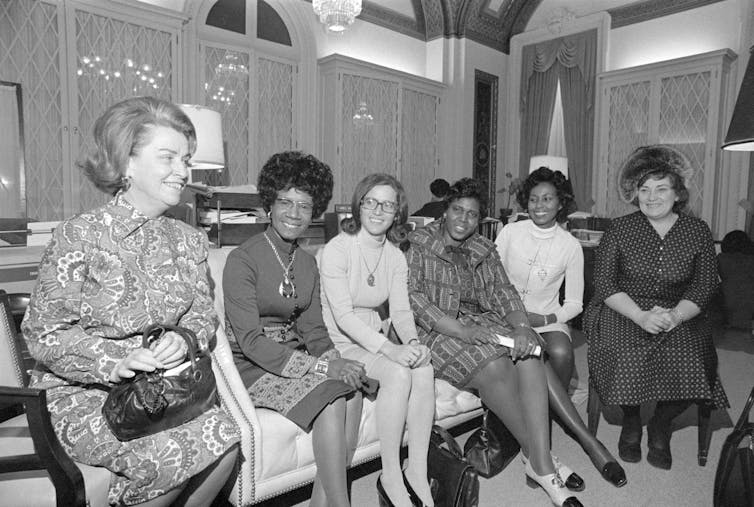 A black and white photo shows four middle-aged women, three Black and three white, in old fashioned formal clothing sitting together.