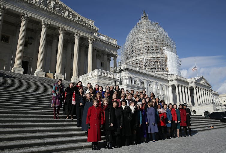 A large group of women wearing jackets stand on a flight of outdoor steps, with two white buildings with pillars behind them.