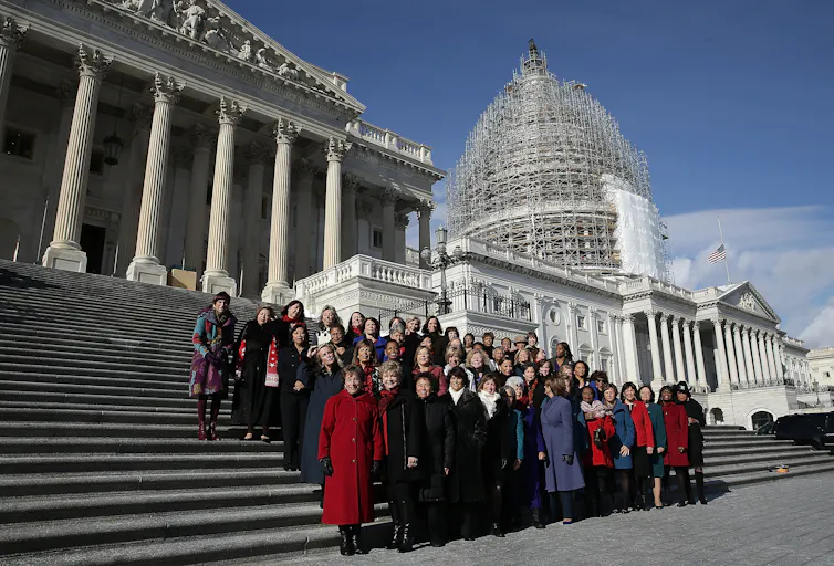 A large group of women wearing jackets stand on a flight of outdoor steps, with two white buildings with pillars behind them.