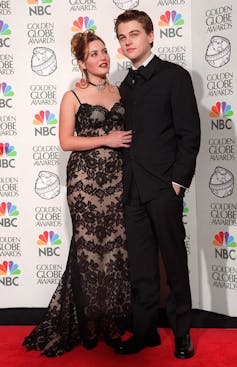 Kate Winslet and Leonardo DiCaprio stand arm in arm on the red carpet. He's in a black tuxedo and she wears a lace black dress.