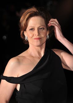 Sigourney Weaver on a red carpet in a long black off shoulder gown, her hair pinned back.