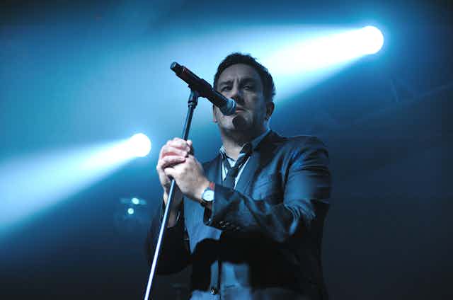 A man, Terry Hall of The Specials, standing in front of a mic on stage beneath a spotlight.