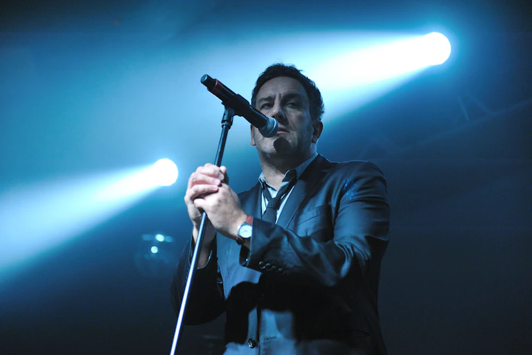 A man, Terry Hall of The Specials, standing in front of a mic on stage beneath a spotlight.