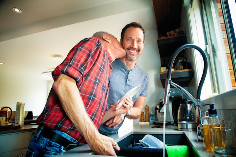 middle-aged man kisses neck of male partner while he is washing dishes at kitchen sink