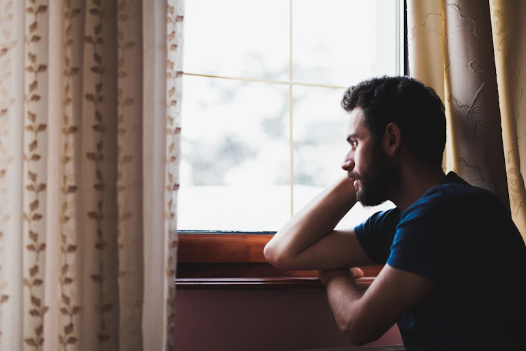 A man wearing a blue shirt looks out of a window with his arm resting on the window ledge.