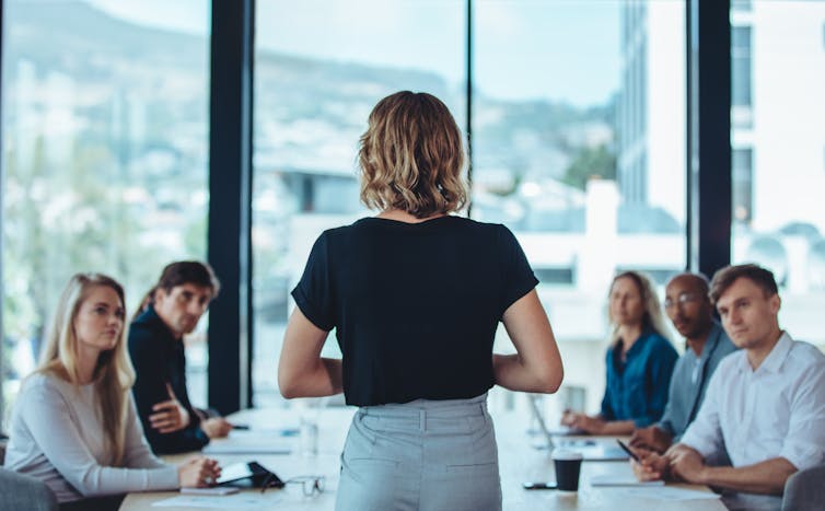 A woman stands with her back to the camera facing a conference table full of people