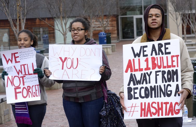 three young people stand in a plaza holding handwritten protest signs