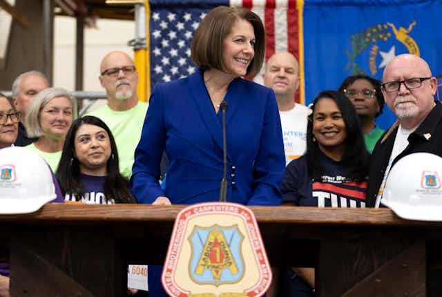 A woman in a blue blazer stands at a lectern with people arrayed behind her.