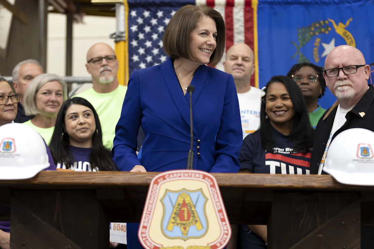 A woman in a blue blazer stands at a lectern with people arrayed behind her.