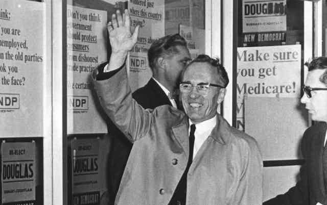 Black and white photo of a smiling man in a raincoat waving with campaign signs in the background, including one reading 'Make sure you get Medicare!'