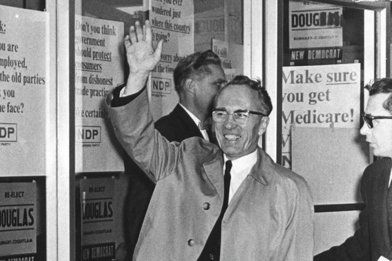 Black and white photo of a smiling man in a raincoat waving with campaign signs in the background, including one reading 'Make sure you get Medicare!'
