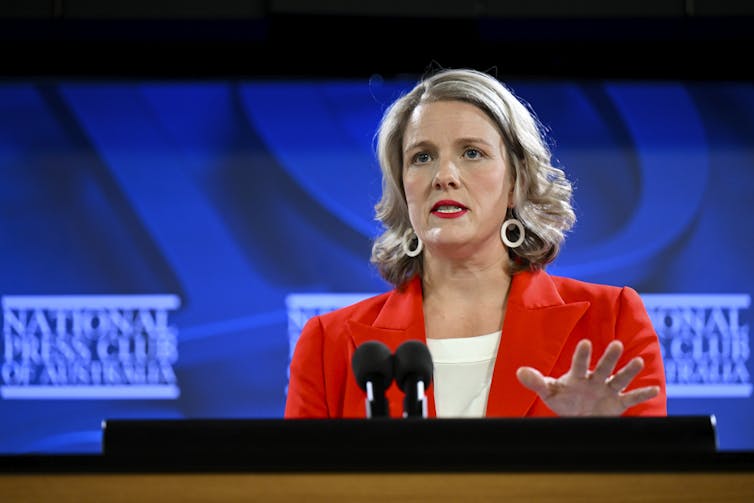 blonde woman in red jacket speaks at lectern