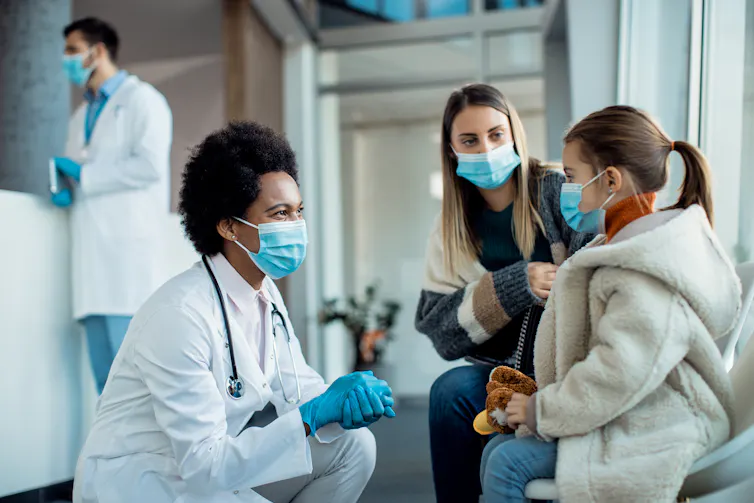 A woman in a white coat, stethoscope and face mask talking to a woman and child in face masks, with a man in a white coat in the background