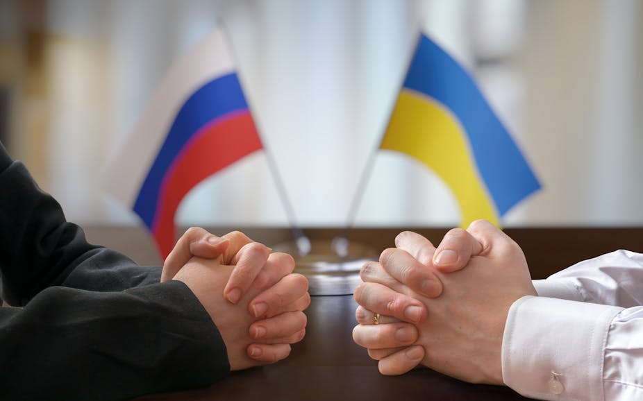 Stock image showing two people's hands clasped across a table with Ukrainian and Russian flags in the background.