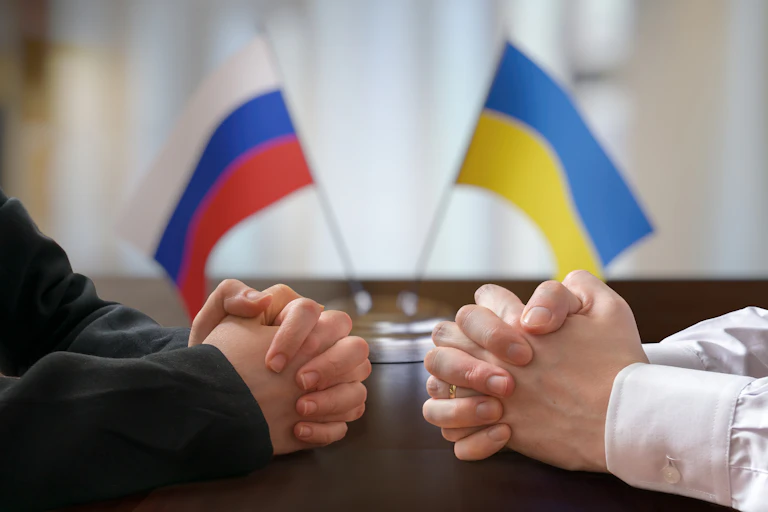 Stock image showing two people's hands clasped across a table with Ukrainian and Russian flags in the background.
